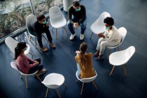 High angle view of group therapy participants sitting in a circle and talking while wearing protective face masks due to COVID-19 pandemic.