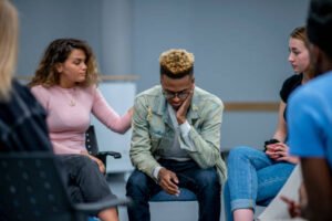 A diverse group of teenagers sit in a group therapy circle and console each other.