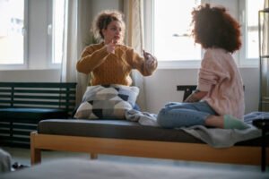 Young woman sitting on the bed with her friend and having a private conversation, explaining and gesturing excitedly