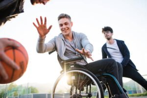 A boy in wheelchair with two teenager friends playing basketball outside at sunset.